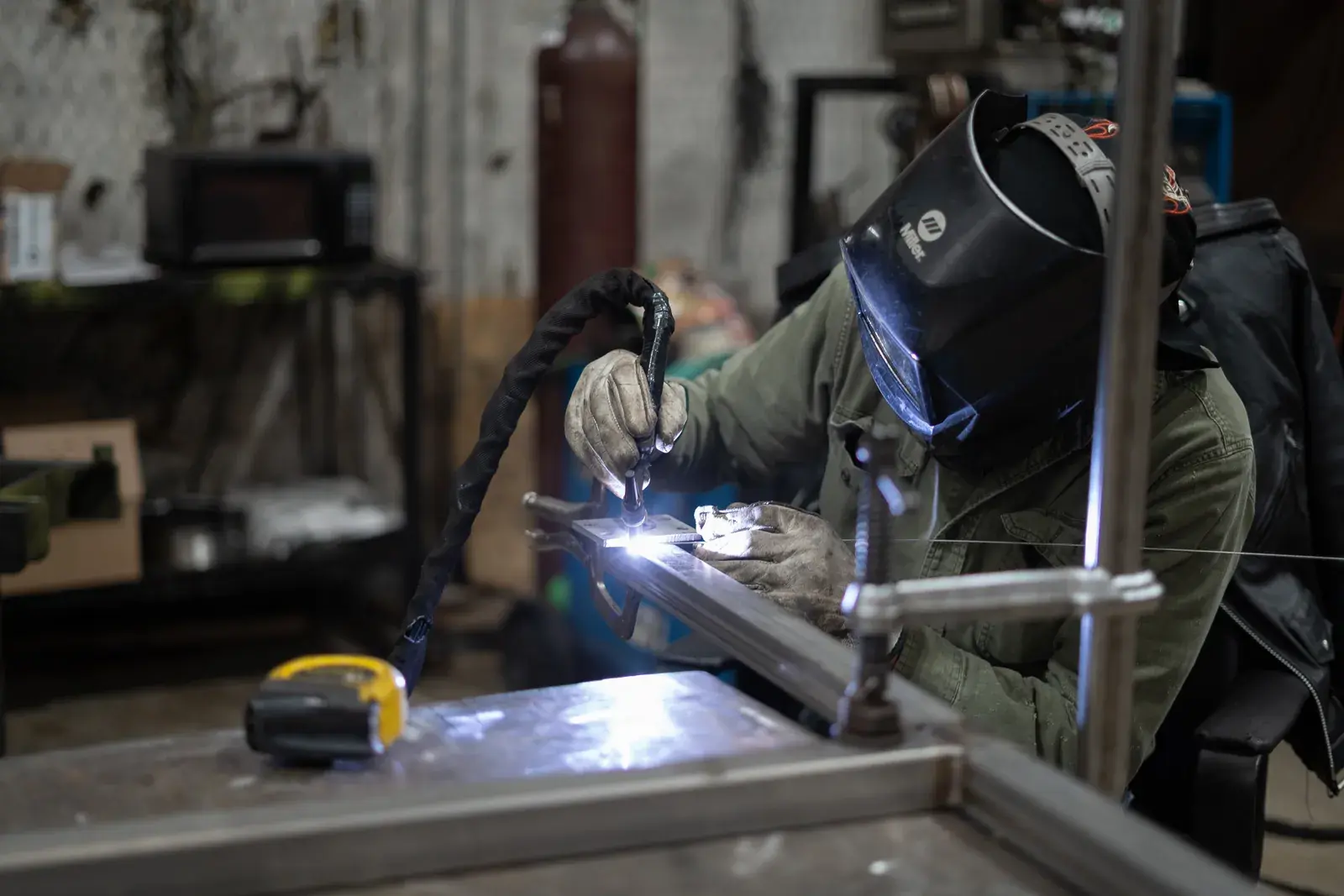 Welder in a helmet, gloves and green jacket welding a clamped metal assembly.