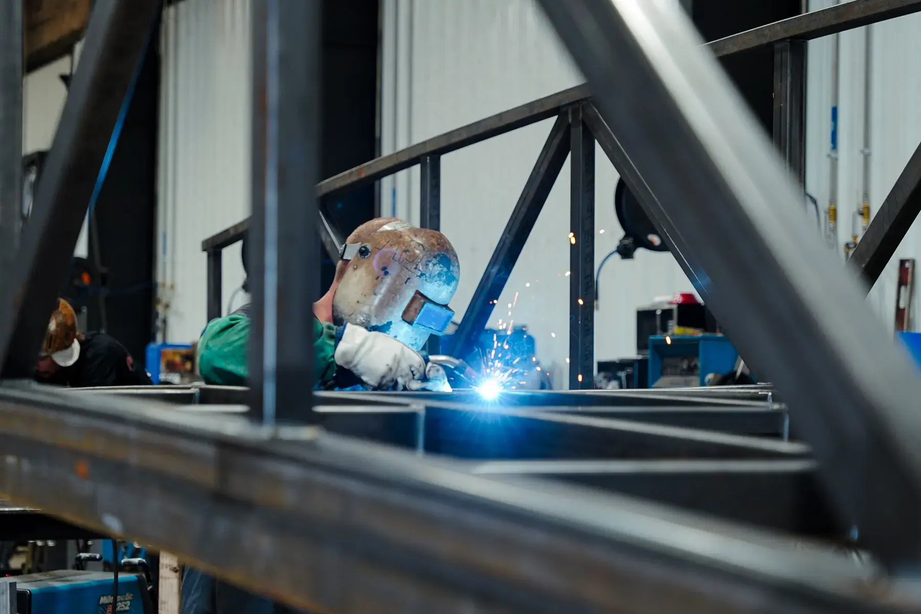 Welder wearing a helmet and gloves, seen through a fabricated steel frame with blue light at the weld.