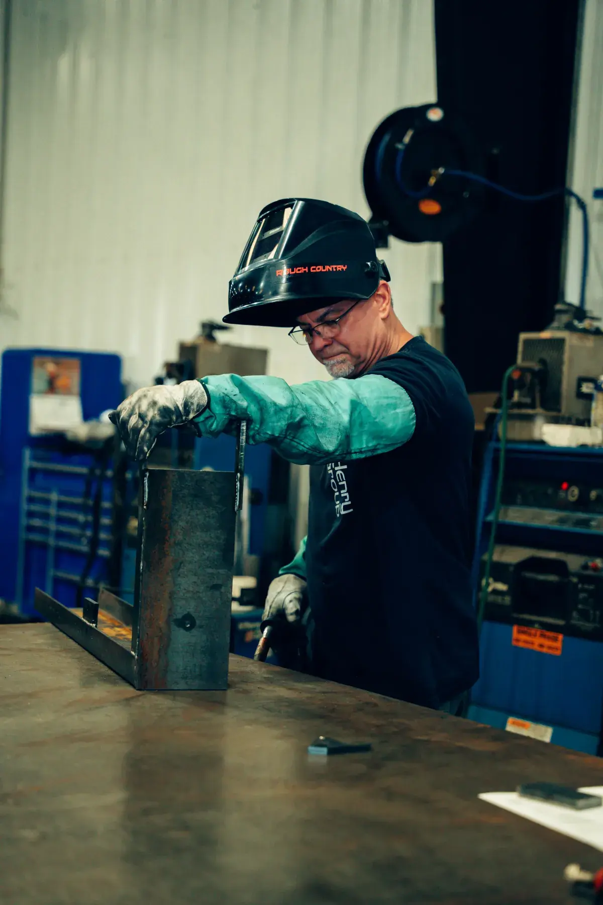 Fabricator wearing gloves and protective sleeves positioning a metal assembly at a workbench, with a welding helmet raised.