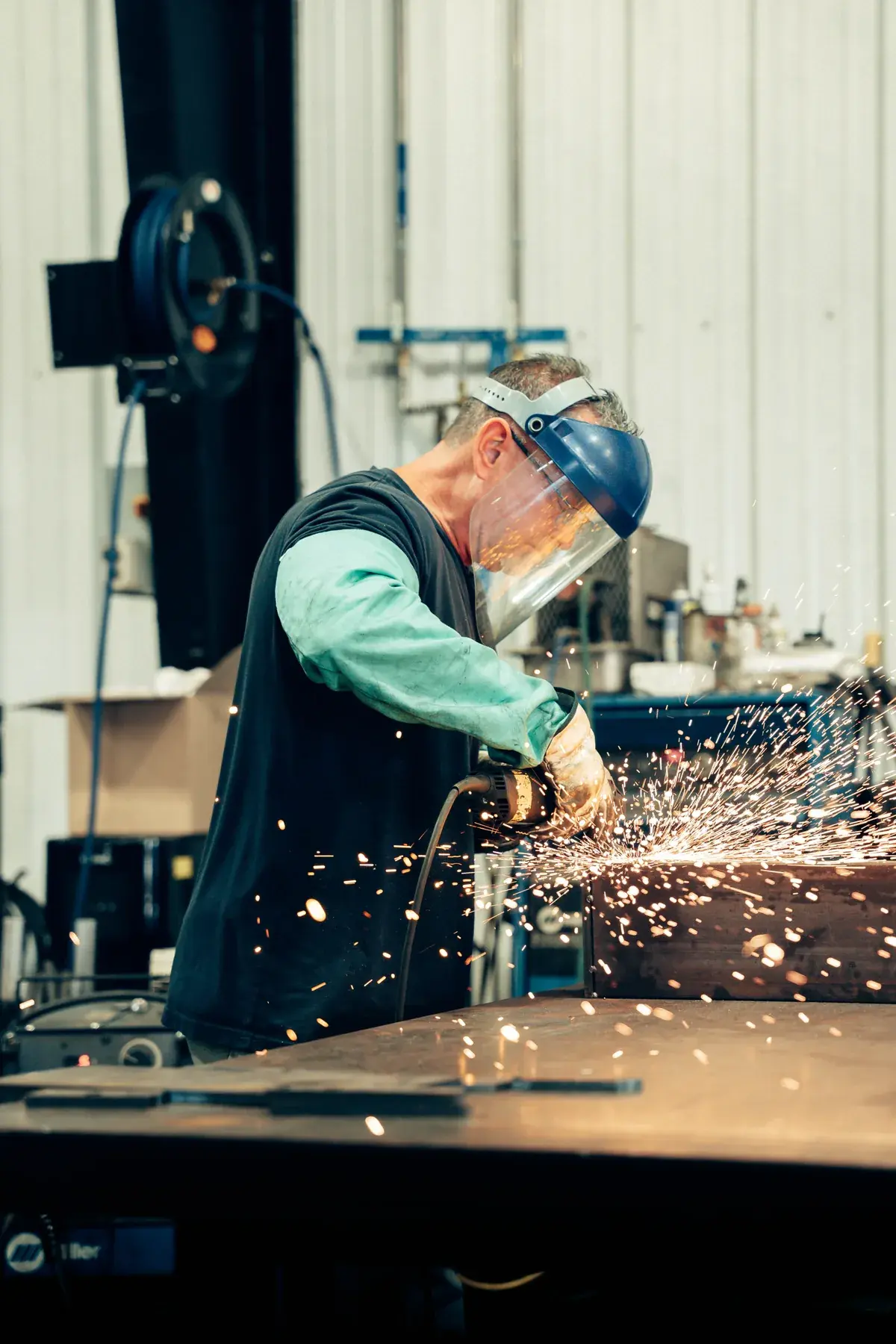 Fabricator wearing a face shield and gloves grinding a metal assembly, with sparks at the tool's contact point.